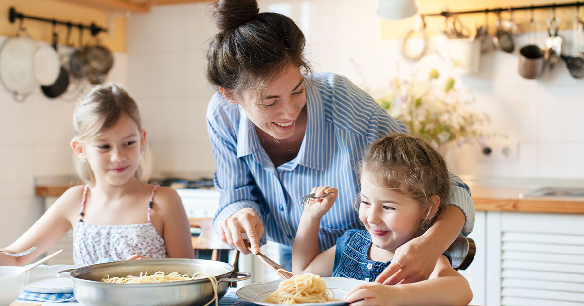 Parent serving pasta to two young children seated at a kitchen table during a family meal.